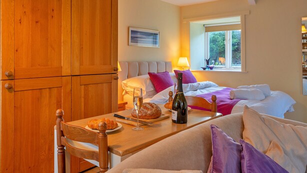 Ein gemütliches Zimmer mit einem Holzschrank, einem Esstisch mit Brot und Wein, einem Bett mit Kissen und einem Fenster mit Blick ins Grüne.