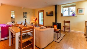 A cozy living room with a fireplace, a dining table with chairs, a sofa, and a bed with a pink blanket. - Studio in Boscastle Near Tintagel Castle (Boscastle)