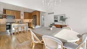 A modern kitchen with wooden cabinets, a central island, bar stools, a dining table with wicker chairs, and a chandelier.
