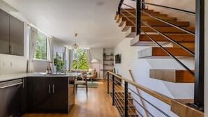 Una cuina moderna amb armaris foscos, una illa de fusta i una zona de dinar amb un sofà i taula. Una escala de fusta amb baranes negres condueix a un pis superior.