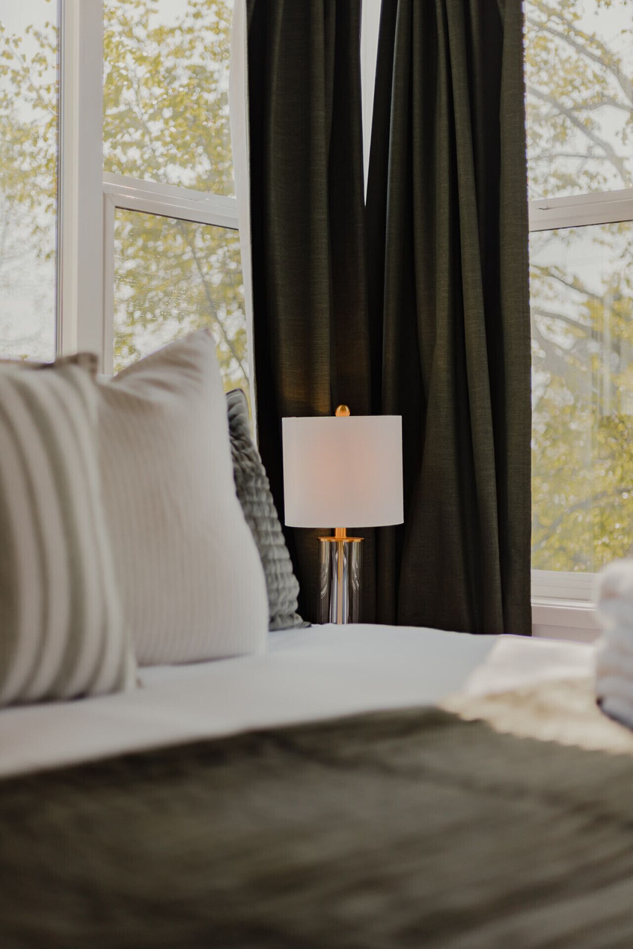 A bed with a white pillow, a grey pillow, and a white blanket. There is a white lamp with a metallic base on a bedside table. The room has a window with dark curtains and a view of trees outside.