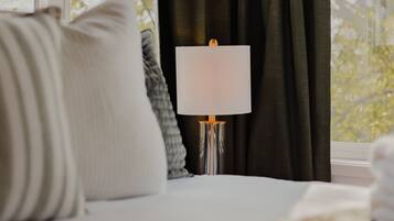 A bed with a white pillow, a grey pillow, and a white blanket. There is a white lamp with a metallic base on a bedside table. The room has a window with dark curtains and a view of trees outside.