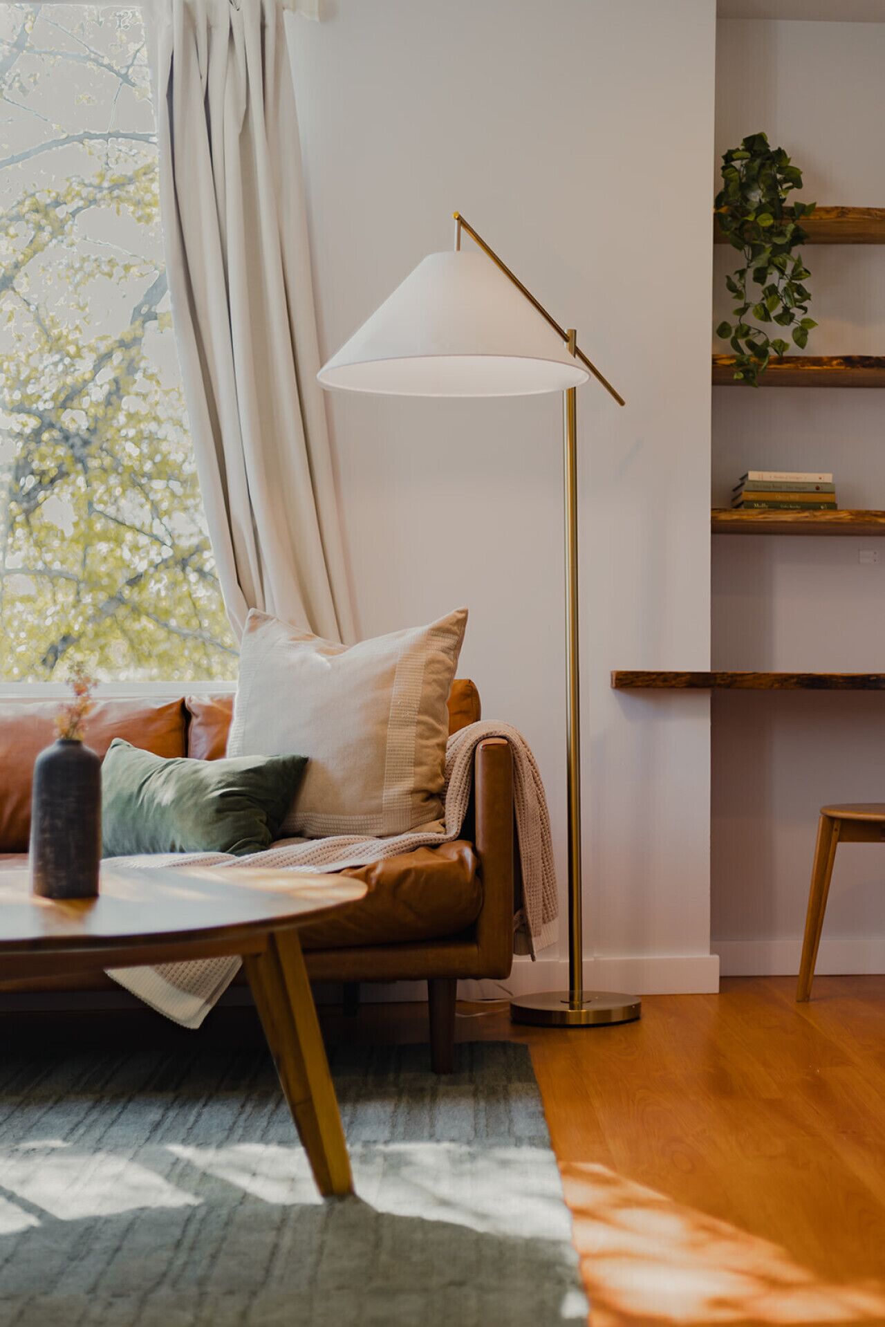 Ein gemütliches Wohnzimmer mit einer Couch, einer Stehlampe, einem Bücherregal und Blick auf Bäume durch das Fenster.