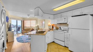 A compact kitchen with white cabinets, a stove, microwave, and a refrigerator. There's a dining area with a table and chairs, and a view of the ocean through large windows.
