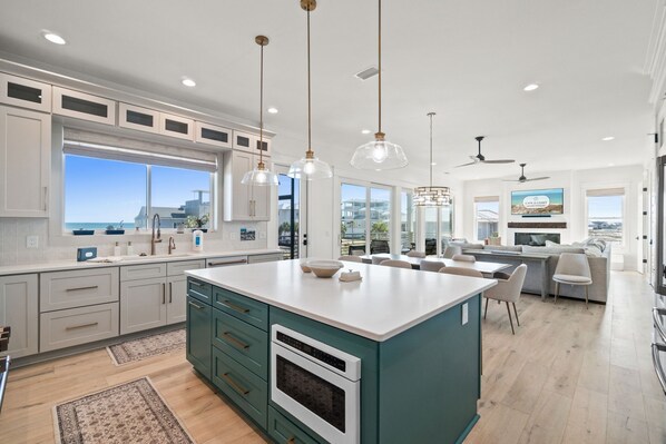 A modern kitchen with white cabinets, a teal island, pendant lights, and a large window offering a view of the city. - Maison Bleu: Beautiful Beach Views, Private Pool, Pet Friendly, East End (St. George Island)