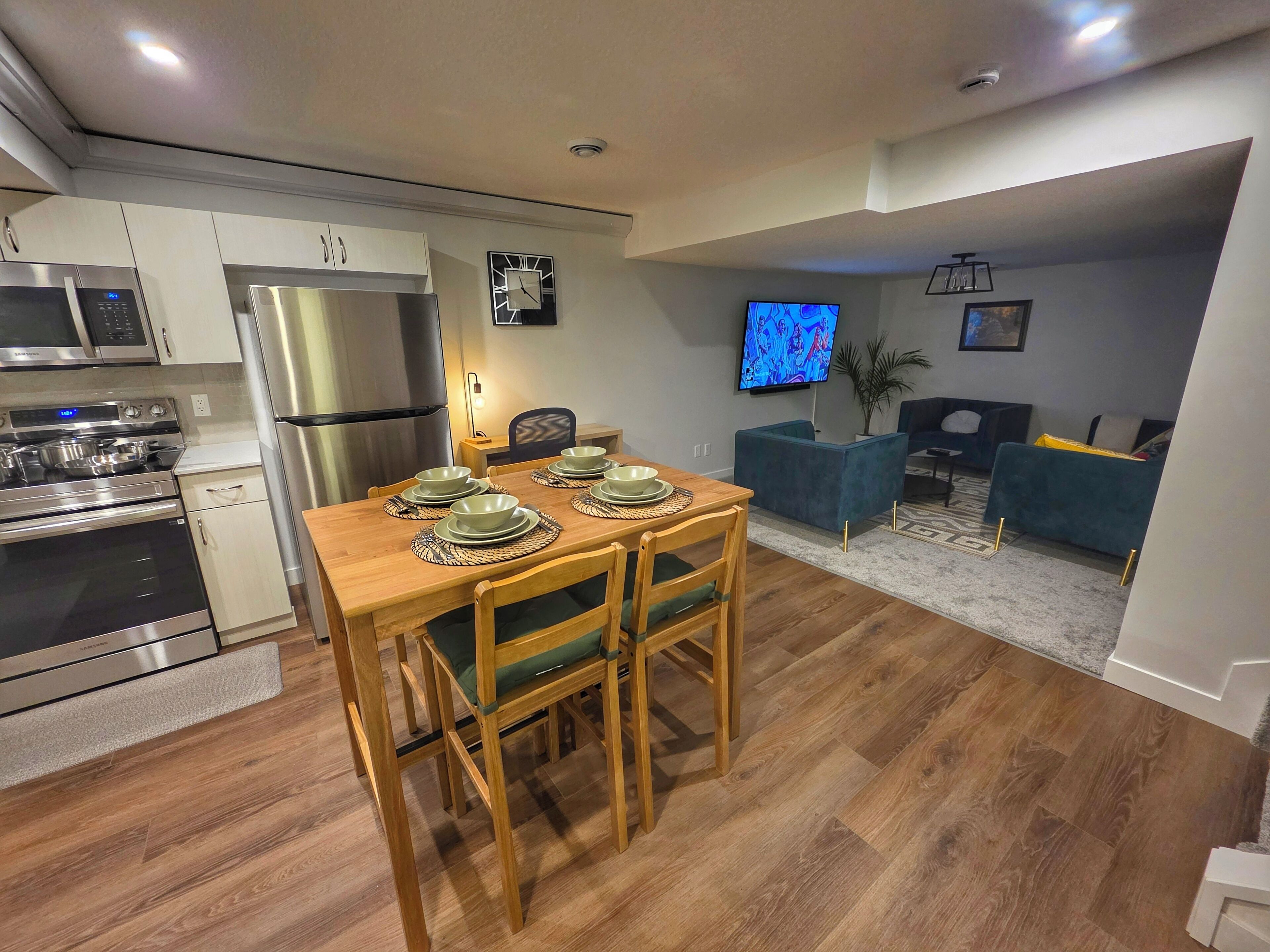 A modern kitchen with stainless steel appliances, a wooden dining table set for four, and a living area with a sofa and TV.