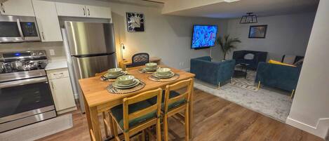 A modern kitchen with stainless steel appliances, a wooden dining table set for four, and a living area with a sofa and TV.