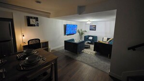 Una sala de estar moderna con un televisor de pantalla plana, sofás oscuros, una mesa de comedor para cuatro personas y una mesita auxiliar.