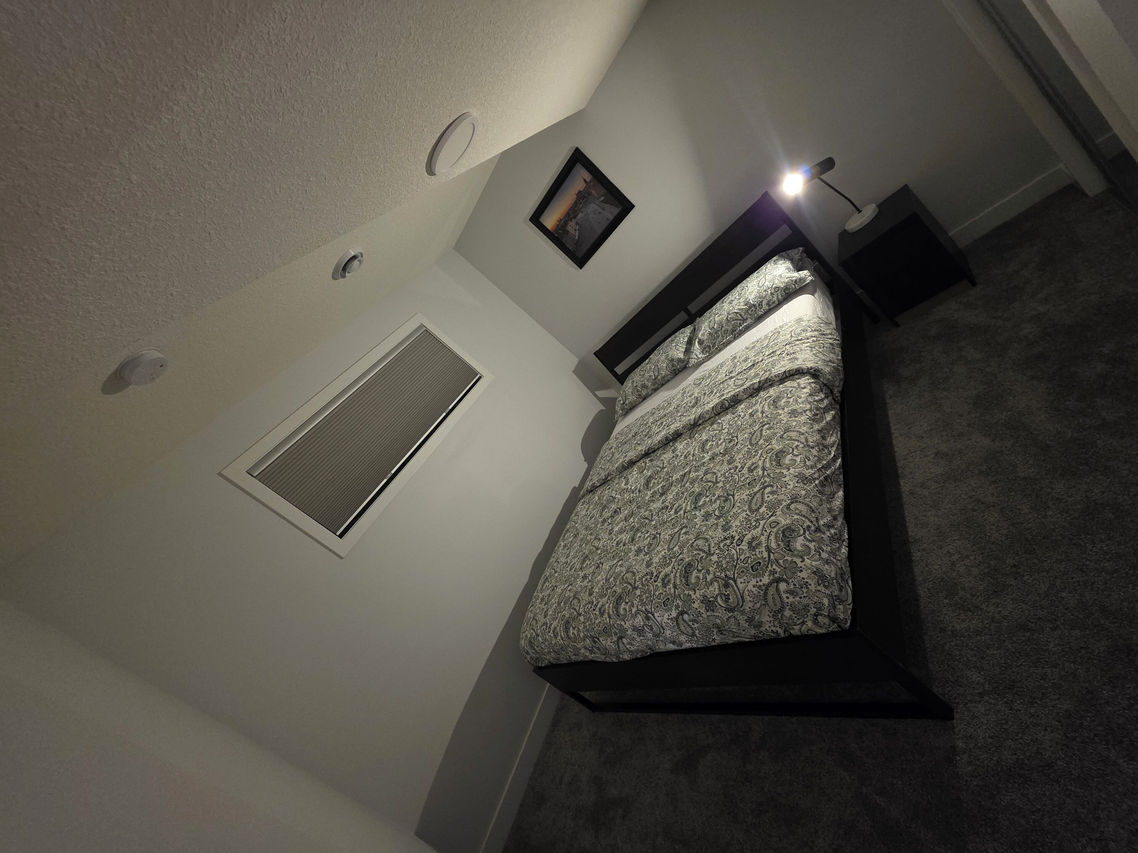 Una camera da letto con un letto, un comodino, una lampada, un quadro appeso al muro e una bocchetta di ventilazione.