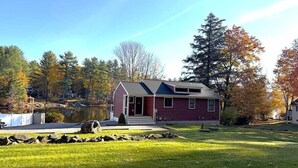 A single-story house with a red exterior and a grey roof, surrounded by a well-maintained lawn and a small pond. - Lakeside Bliss at Pickerel Cove (Laconia)