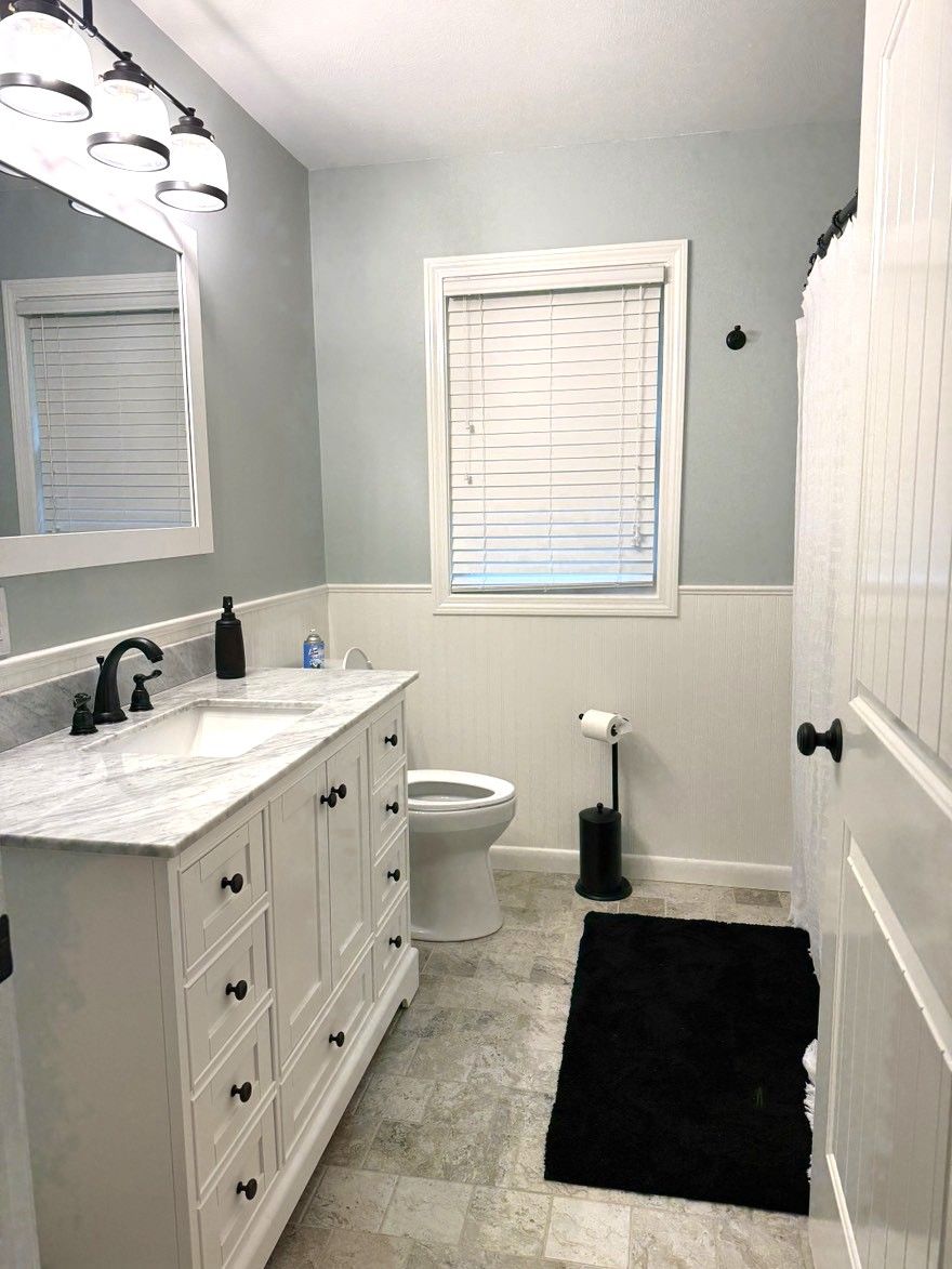 A bathroom with a white vanity, a toilet, a window with blinds, and a black rug.