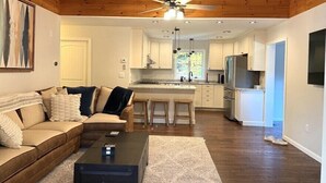 A modern living room with a wooden ceiling, a ceiling fan, a sofa, a coffee table, and a kitchen area with white cabinets and a refrigerator. - Lakeside Bliss at Pickerel Cove (Laconia)