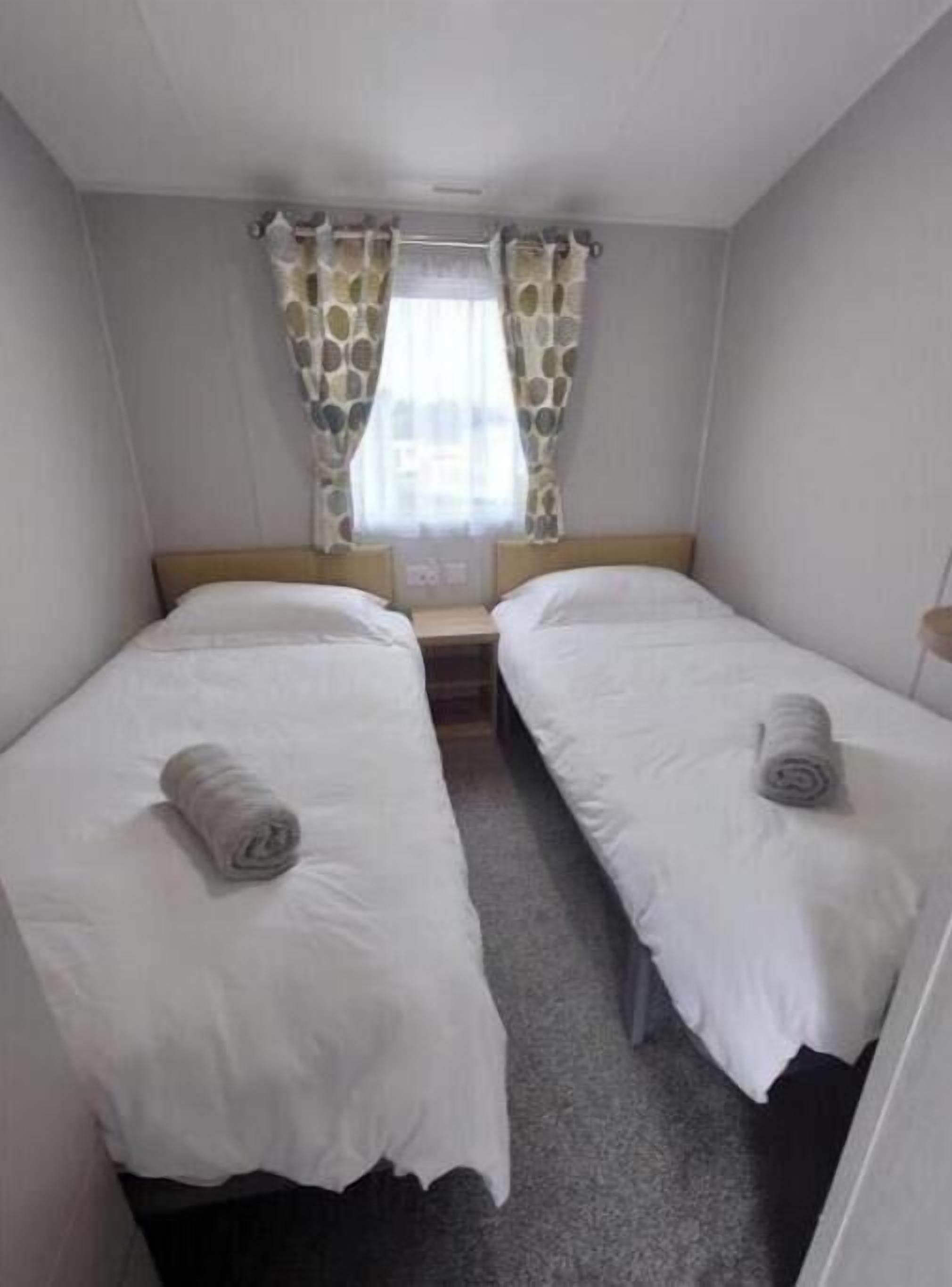 Une petite chambre propre, avec deux lits simples, une fenêtre ornée de rideaux à motifs et un tapis uni.