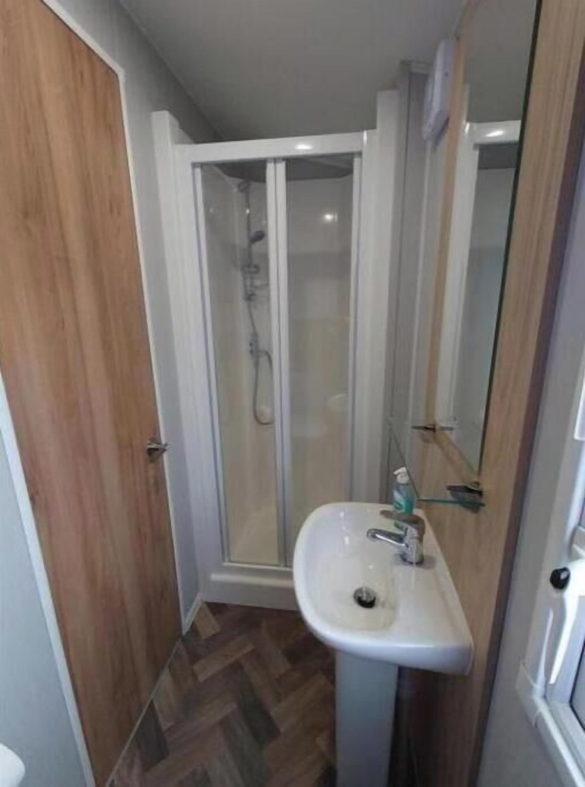 Une salle de bain avec une porte en bois, une douche en verre et un lavabo blanc.