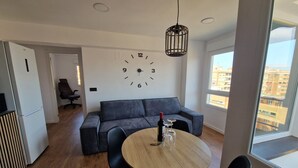 Un salon moderne avec une table ronde en bois, des chaises noires, un canapé gris, une horloge murale et une grande fenêtre donnant sur la ville.
