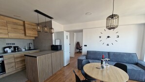 Une cuisine moderne avec des armoires en bois, un îlot central intégré et des luminaires suspendus. Une grande horloge murale et un canapé sont visibles dans l’espace de vie.
