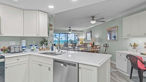 A kitchen with white cabinetry, a dishwasher, and a dining area with a chair and table. - 3rd-Floor Island Getaway w/ Lagoon Views – Walk to the Beach! (Sanibel)