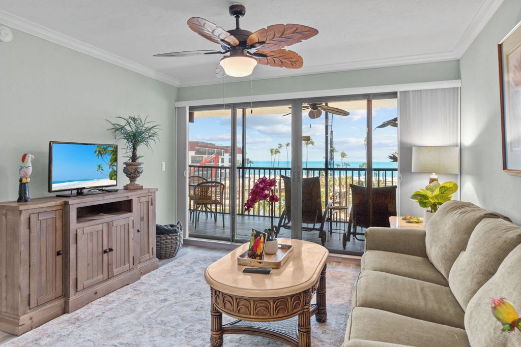 A living room with a television, a wooden cabinet, a round coffee table, a beige sofa, and a balcony with outdoor furniture and a view of the ocean.