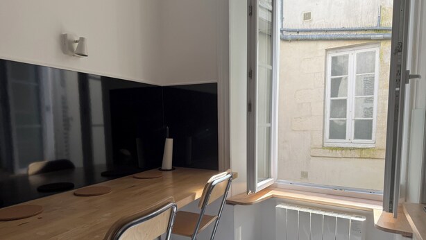 A modern kitchen with a wooden countertop, two metal chairs, a white wall, and a window with a view of a building outside. - Charming bright studio (La Rochelle)