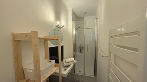 A compact bathroom with a wooden shelf unit, a mirror, a towel rack, and a glass shower enclosure. - Charming bright studio (La Rochelle)