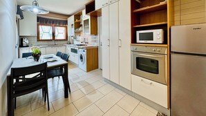 Een kleine keuken met witte kastjes, een ingebouwde magnetron, een fornuis en een spoelbak. Er is een eettafel met stoelen en een hangende lamp.