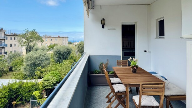 Een balkon met een houten tafel en stoelen, een bloempot en uitzicht op gebouwen en groen.