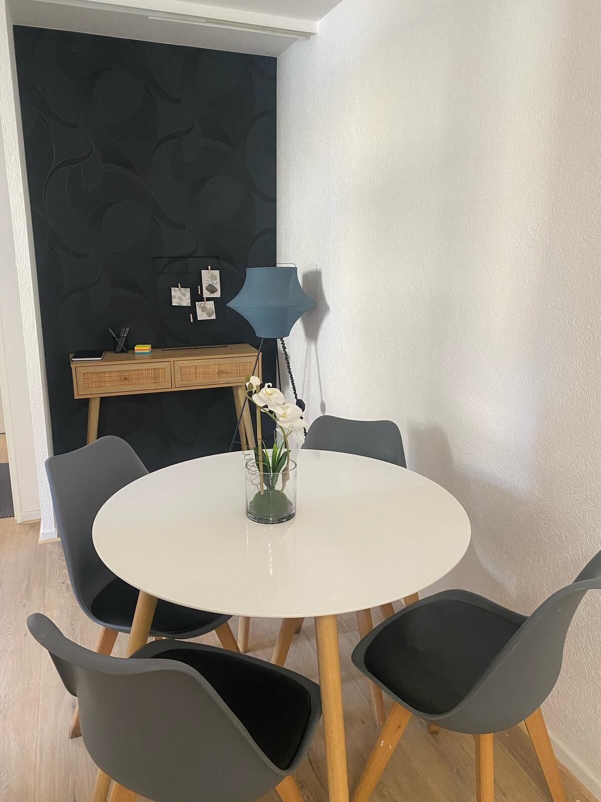 A round white table with four black chairs, a small vase with white flowers, and a blue lamp on a wooden cabinet against a dark patterned wall.