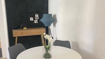 A round white table with four black chairs, a small vase with white flowers, and a blue lamp on a wooden cabinet against a dark patterned wall.