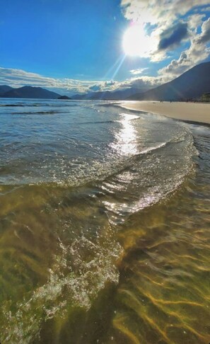 Een strand met helder ondiep water, een zandstrand en verre bergen onder een licht bewolkte lucht.