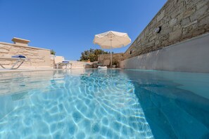 Piscina cristalina con sillones de descanso y sombrillas, paredes de piedra y un cielo azul despejado.