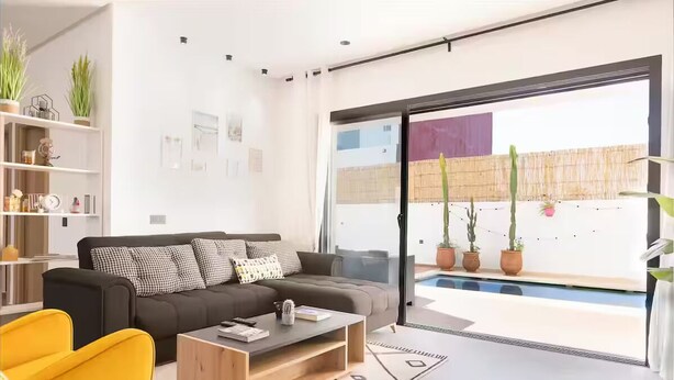 Una sala de estar moderna con un sofá, una mesa de centro y un sillón amarillo. Hay plantas en macetas y un ventanal con vistas a una piscina y zona exterior.