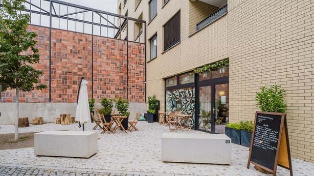 Un espace extérieur aménagé avec des tables et des chaises, un tableau noir servant de menu, et un bâtiment en briques en arrière-plan.