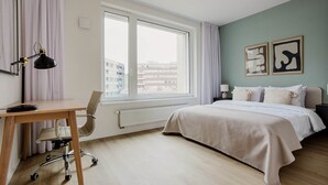 Une chambre moderne dotée d’une grande fenêtre, d’un bureau avec une chaise, d’un lit recouvert d’une couette blanche et de deux œuvres d’art abstraites accrochées au mur.