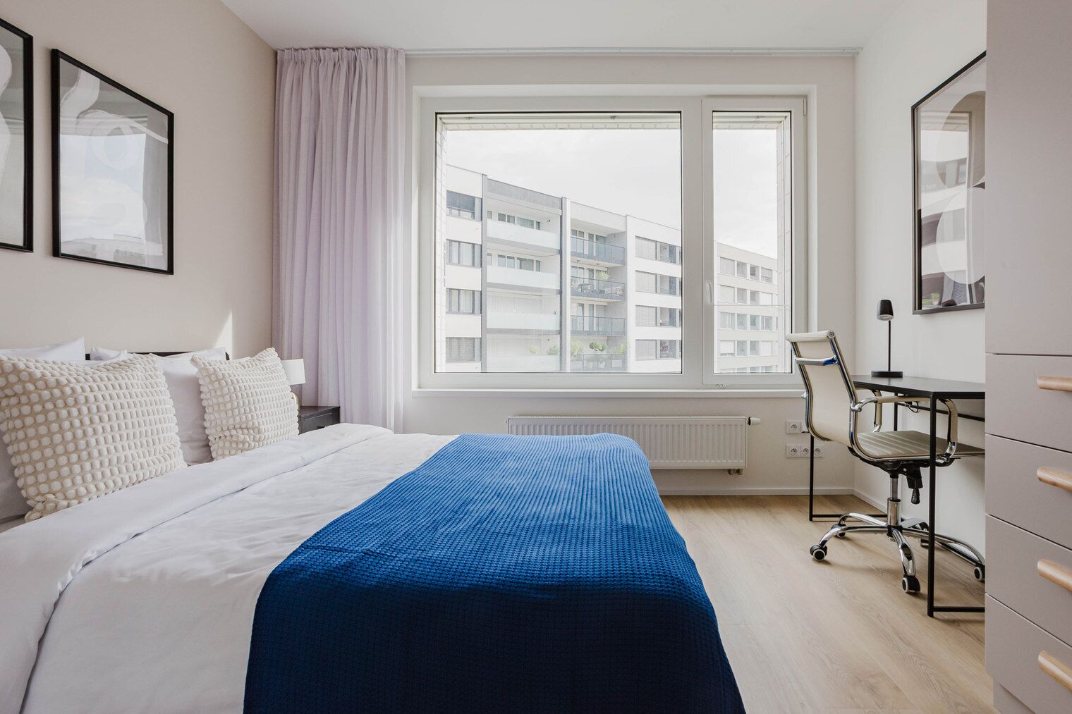 Ein modernes Hotelzimmer mit einem großen Bett, einem Schreibtisch mit Stuhl und Blick auf die Stadt durch das Fenster.