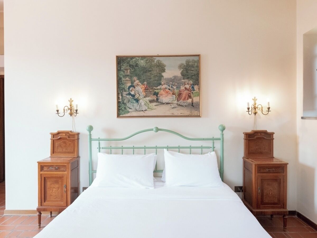 Un dormitorio con estructura de cama metálica verde, ropa de cama blanca y dos mesitas de noche de madera con lámparas de pared.
