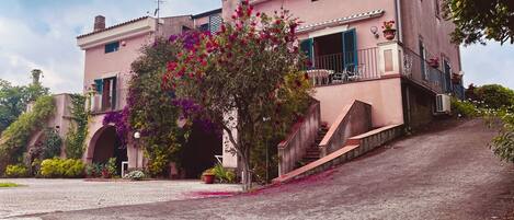A two-story building with a pink facade, a balcony with blue shutters, and a staircase leading to the entrance.