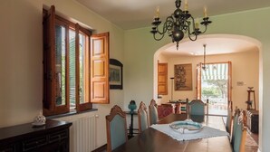 Comedor con gran mesa de madera, sillas y una lámpara de araña. Se ve una ventana con contraventanas de madera y vistas a follaje exterior.