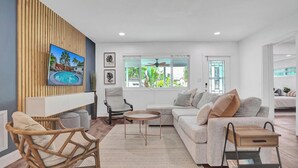 Uma sala de estar moderna com vista para a piscina, um sofá, uma poltrona e uma TV fixada numa parede de madeira.
