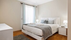 A bedroom with a bed, bedside tables, a nightstand, a window with curtains, and a wooden floor.