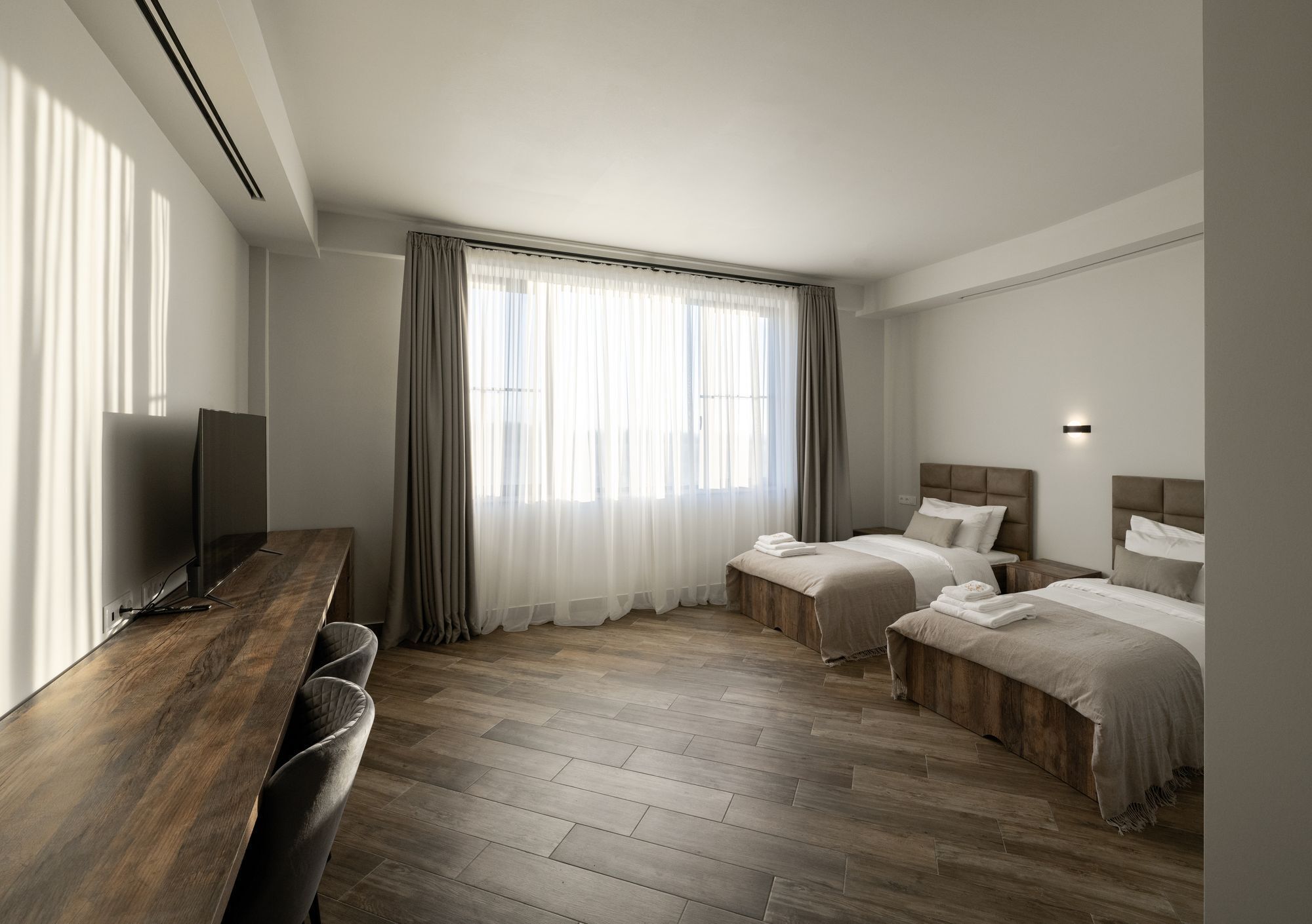 Une chambre d’hôtel moderne avec un plancher en bois, deux lits simples, un bureau avec un téléviseur à écran plat et une grande fenêtre avec des rideaux légers.