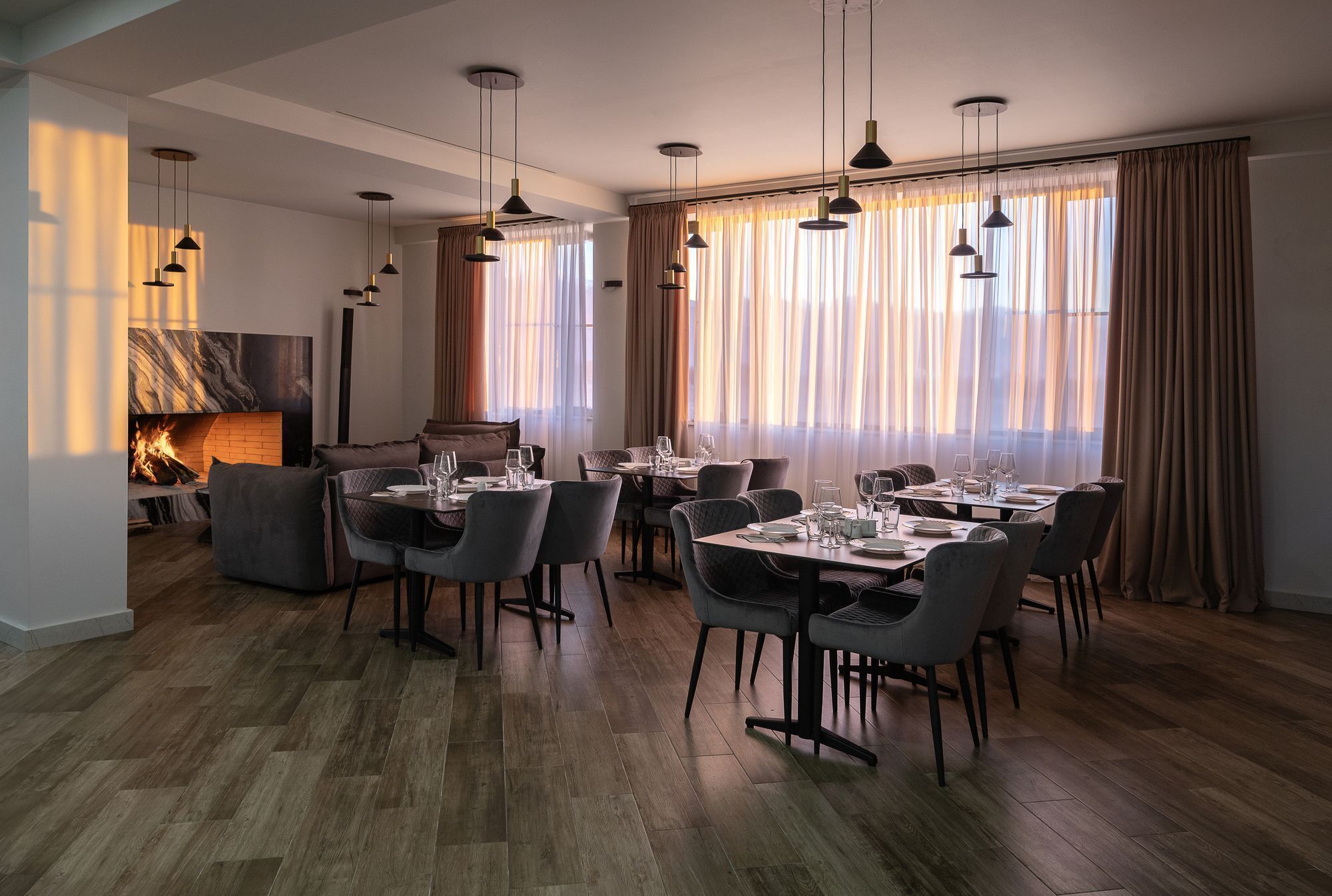 A modern dining area with wooden flooring, multiple tables set for dining, and hanging pendant lights.