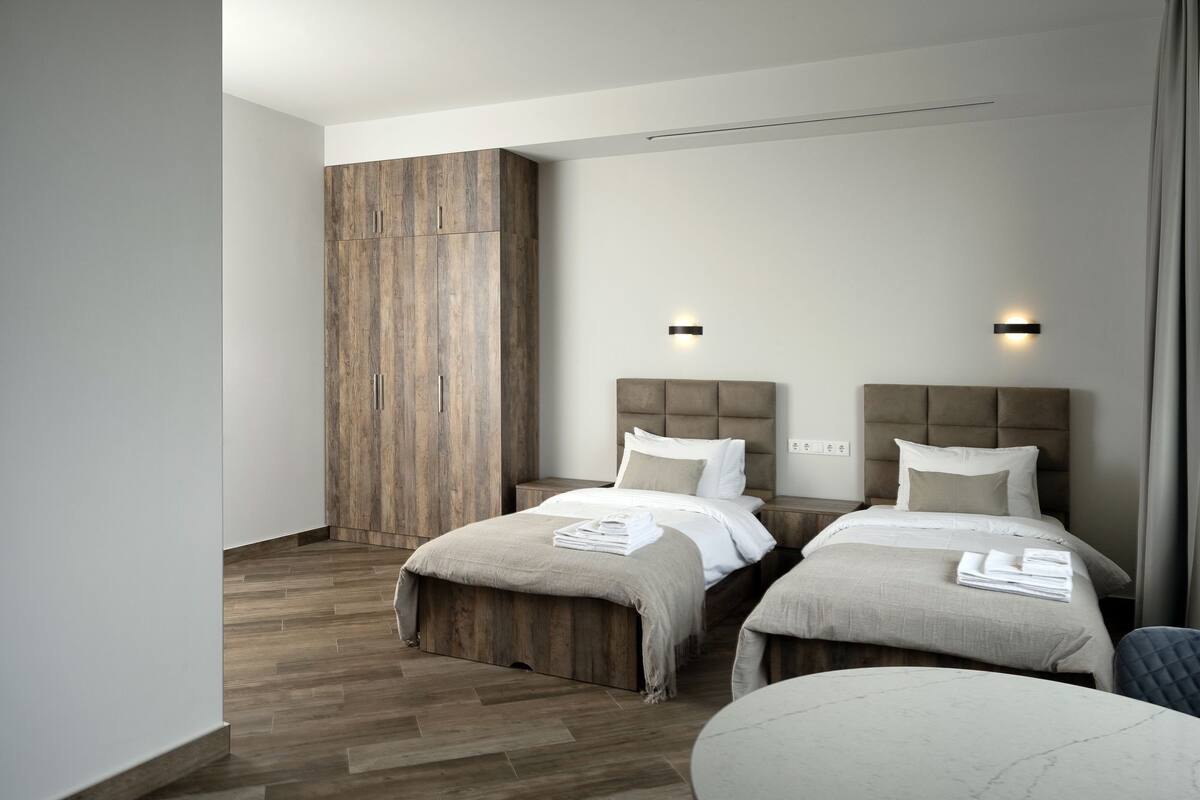 Quarto de hotel moderno com duas camas, um guarda-roupa de madeira, uma mesa redonda e uma janela grande.