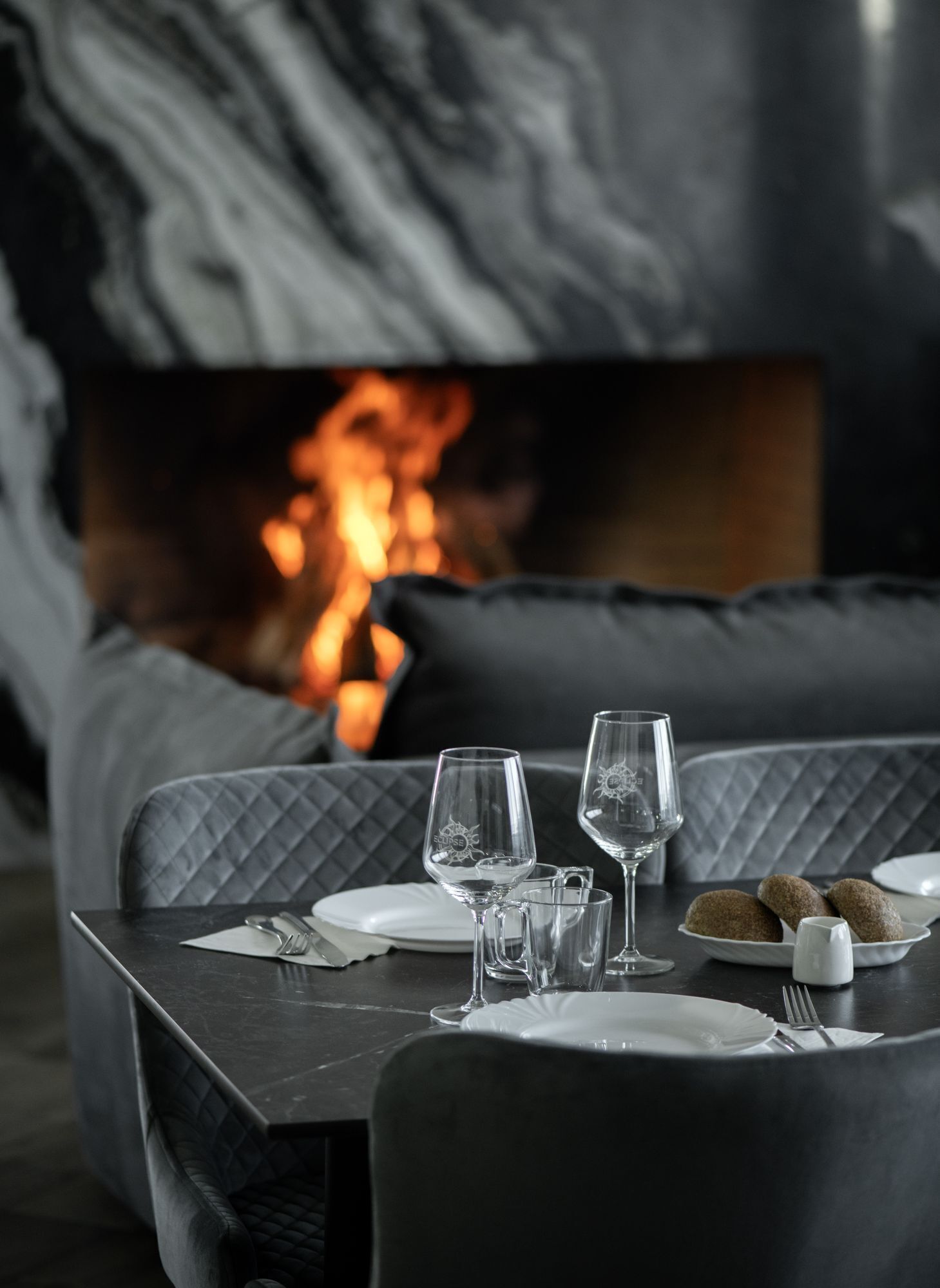 A dining table set with wine glasses, plates, and cutlery, with a fireplace in the background.