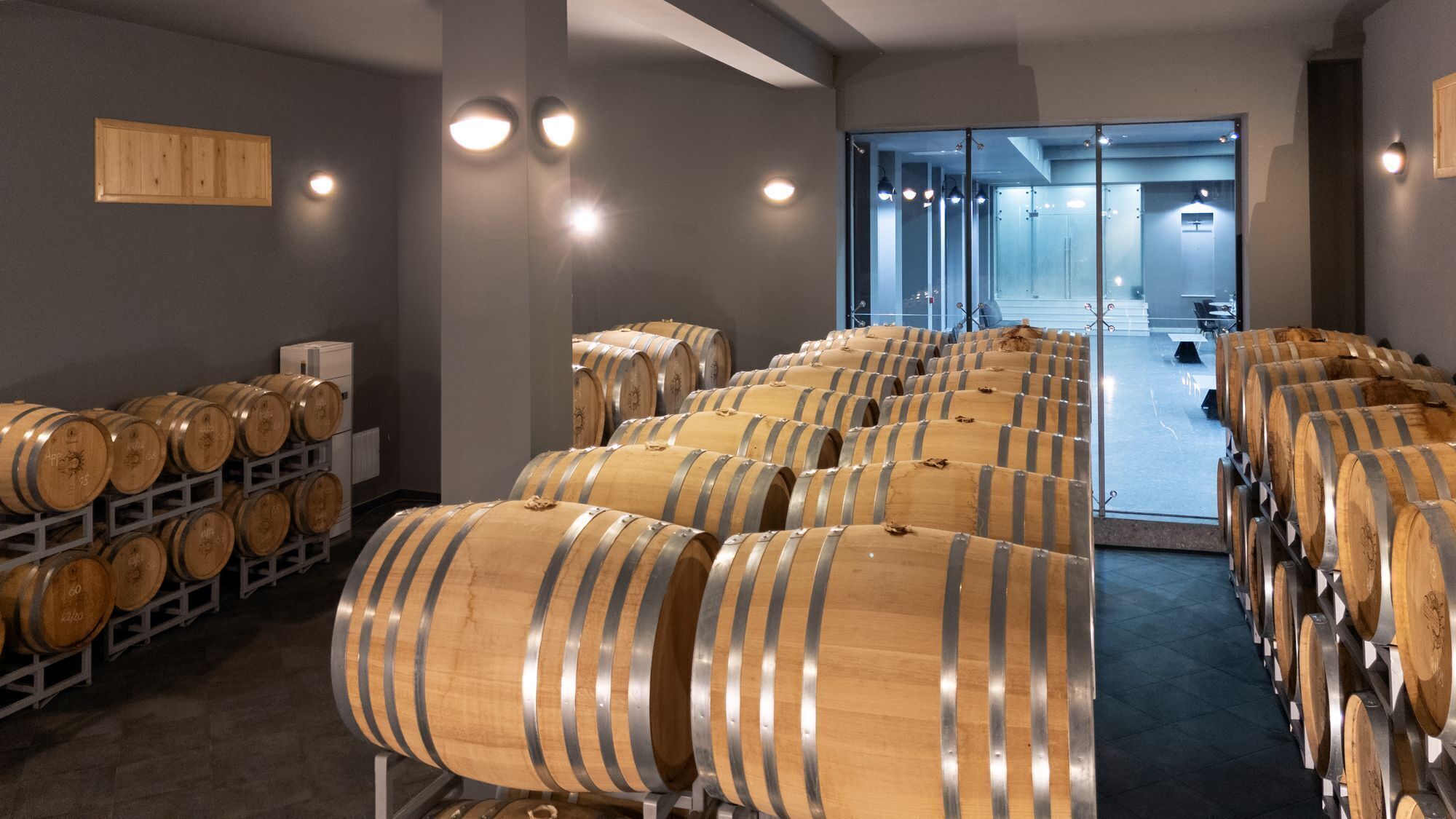 A wine cellar with rows of wooden barrels, a glass door leading to a tasting room, and a wooden panel on the wall.