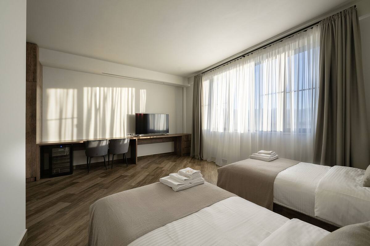 Quarto de hotel com duas camas, uma escrivaninha com televisão e uma janela grande com cortinas.