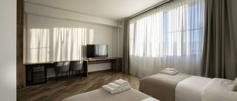 Habitación de hotel con dos camas, un escritorio con televisor y un ventanal con cortinas.