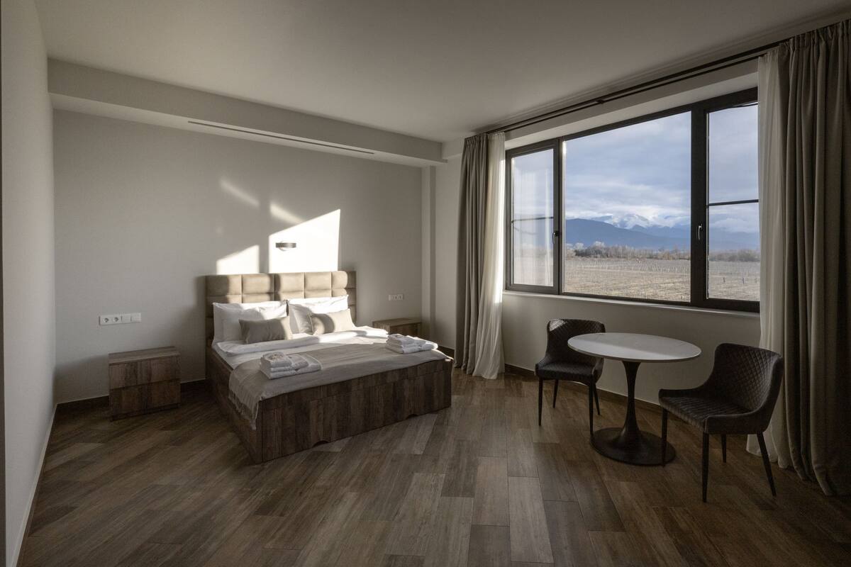 Quarto de hotel moderno com uma cama grande, uma mesinha, duas cadeiras e vista para montanhas através de uma janela ampla.
