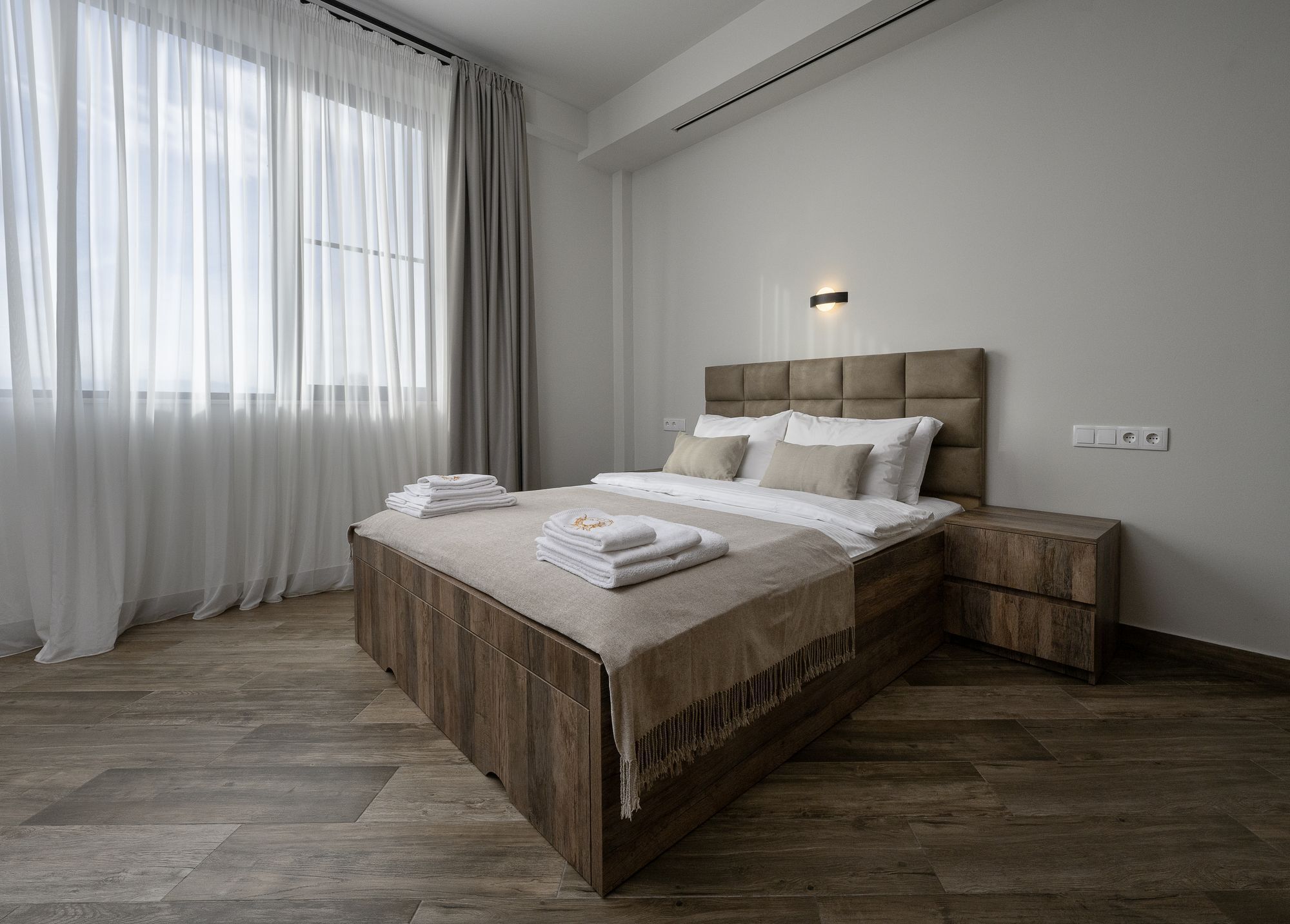 A modern bedroom with a wooden bed, a bedside table, a nightstand, and a large window with sheer curtains.