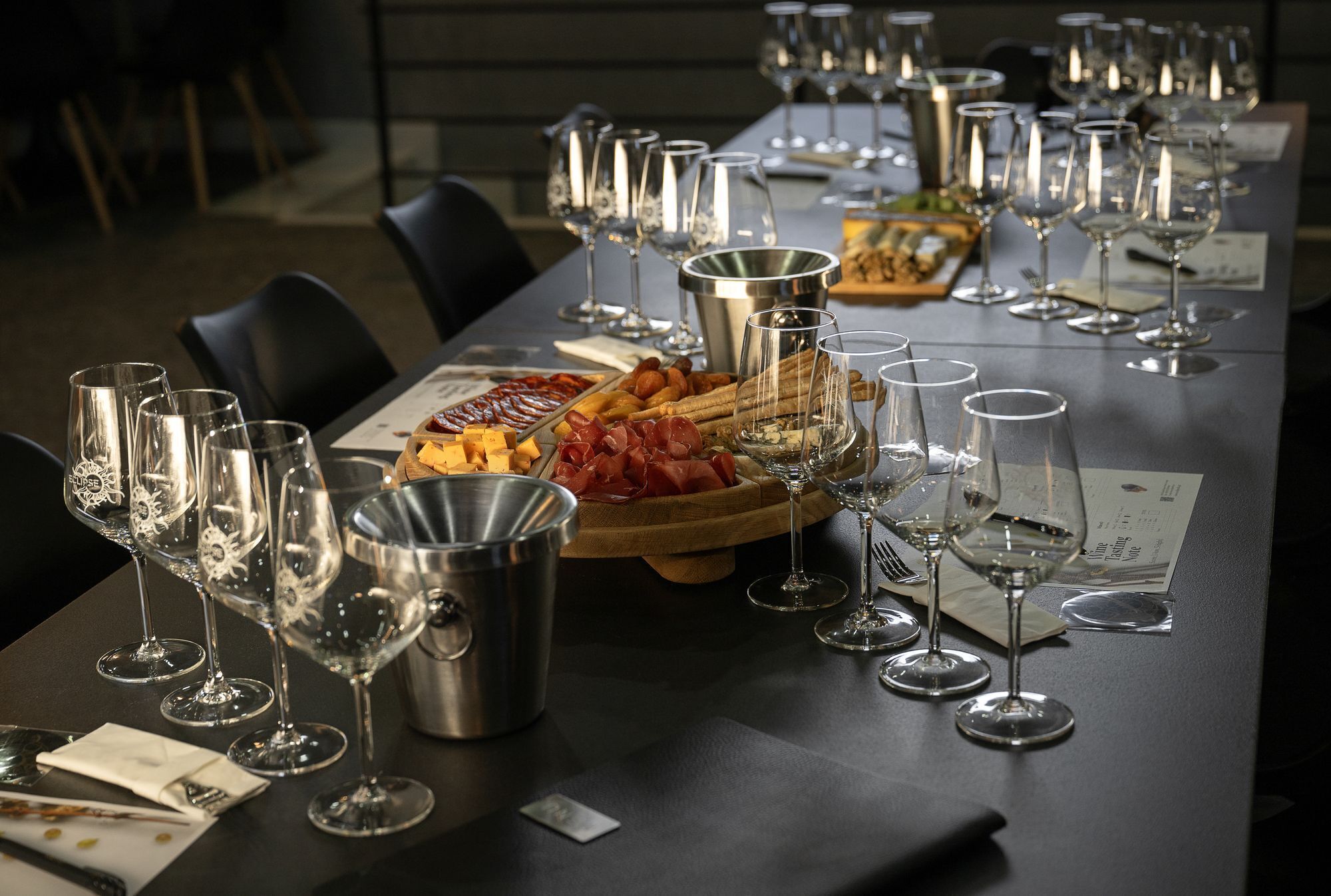 A long table set with wine glasses, a metal bucket, and a wooden board with food items.