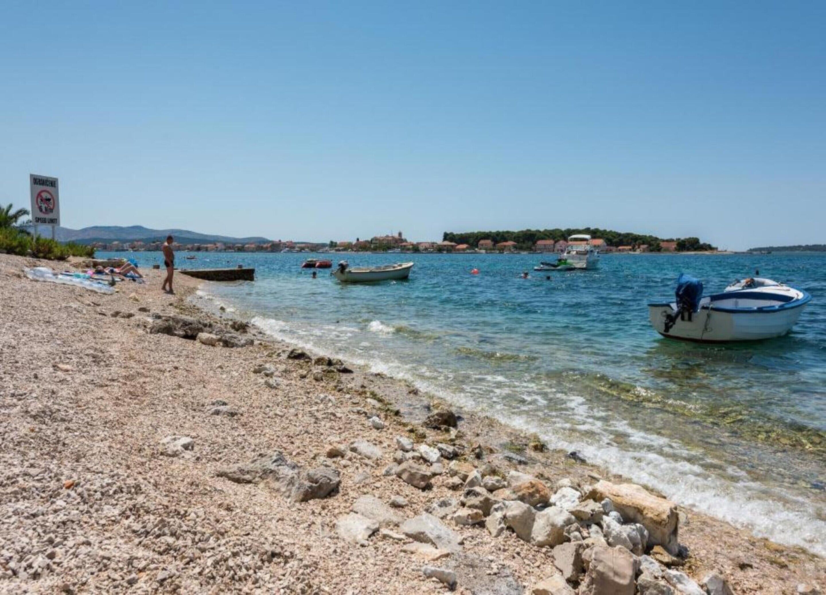 Une plage de galets avec des bateaux, une eau claire et une côte lointaine parsemée de bâtiments.
